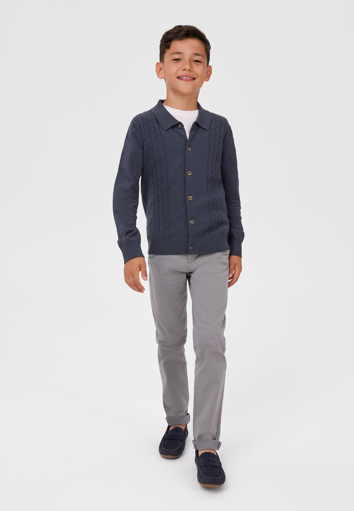 A young boy modeling a navy blue cable-knit button-up cardigan over a white t-shirt, paired with gray trousers and navy loafers, on a light gray studio background.