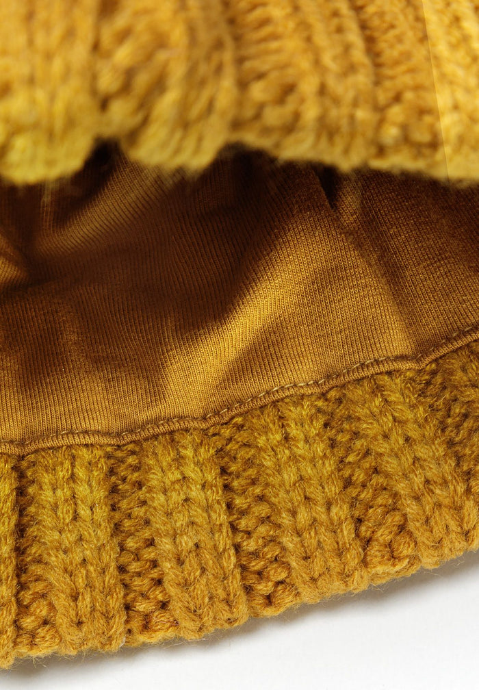 Detail of a mustard knitted cable hat with a pom pom and ribbed edge.