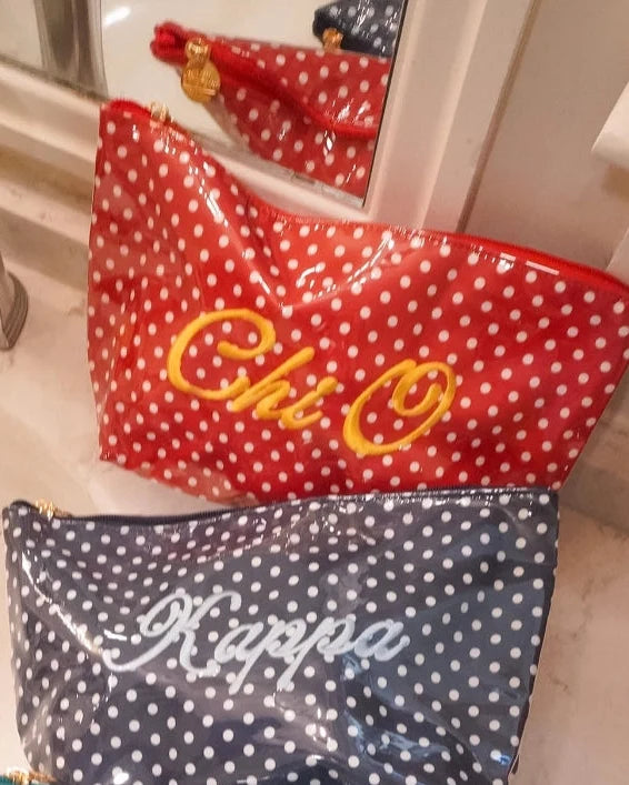Three personalized cosmetic pouches with white polka dots are lined up on a bathroom sink. The top red pouch is embroidered with 'Chi Omega' in yellow, the middle navy blue pouch is embroidered with 'Kappa' in white, and the bottom teal blue pouch is embroidered with 'Zeta' in white.