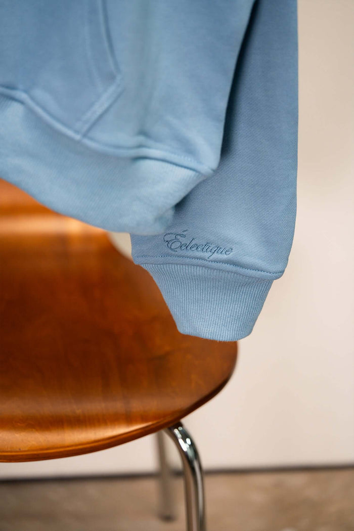 Lightweight terry hoodie with embroidered Éclectique script logo detail on the sleeve, displayed on a wooden chair.