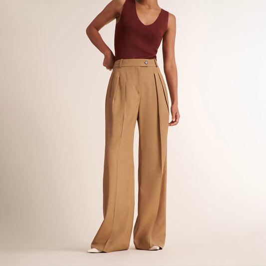 A model wearing high-waisted, wide-leg tan trousers with pleats and a maroon sleeveless top, standing against a plain light beige studio background. | Lead