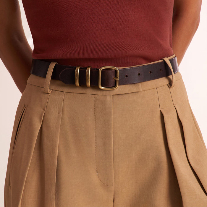A close-up of a person wearing a dark brown leather belt with a gold-tone buckle, styled with pleated tan trousers and a rust-red ribbed top against a light beige background.
