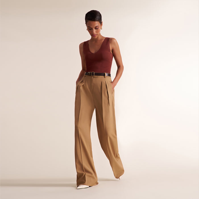A model wearing a sleeveless burgundy top and high-waisted tan wide-leg trousers with a thin black belt, standing against a plain beige studio background.