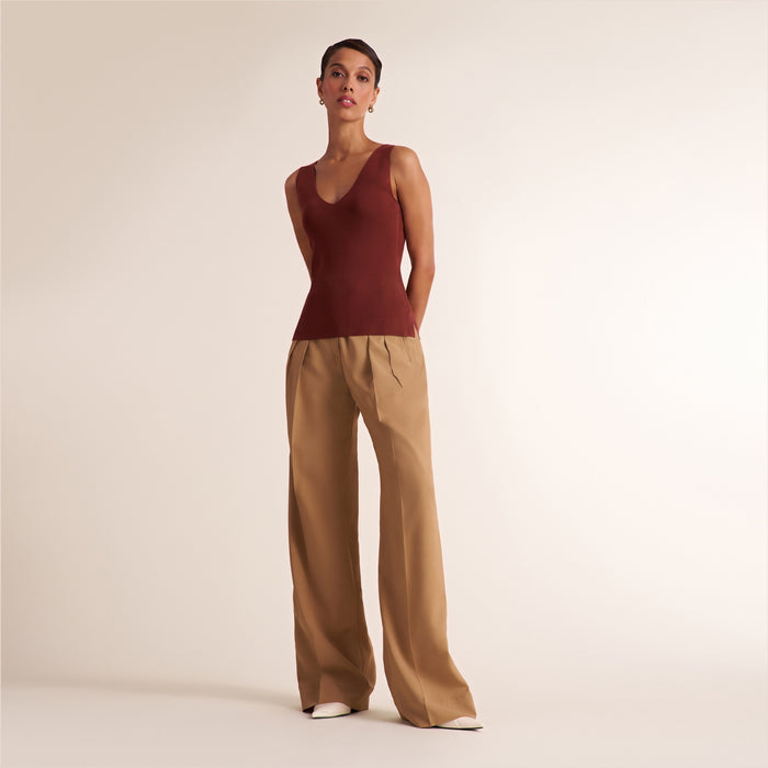 A model stands against a plain light beige studio background wearing a sleeveless deep burgundy V-neck top and wide-leg tan pants.