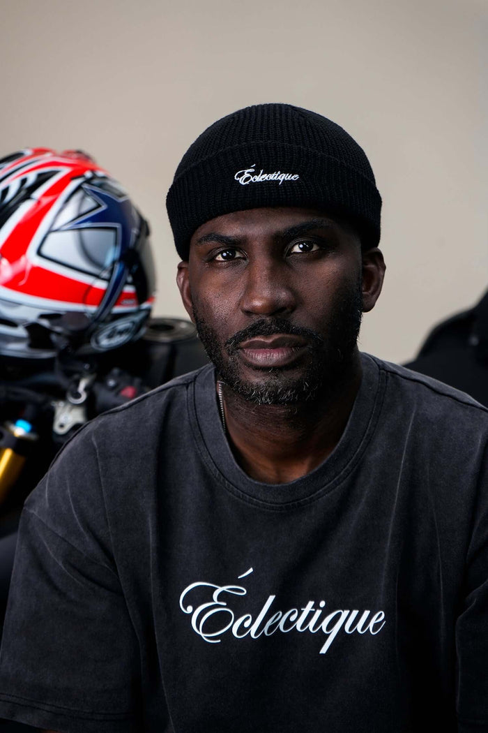 Man wearing a black fisherman beanie, showcasing Éclectique logo, with motorcycle helmet in the background.