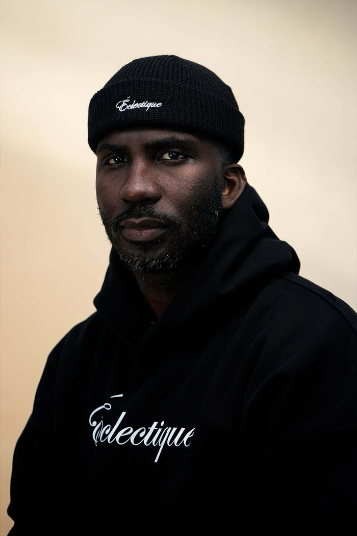 Model wearing a black fisherman beanie and a black hoodie with Éclectique logo, showcasing a stylish urban look.