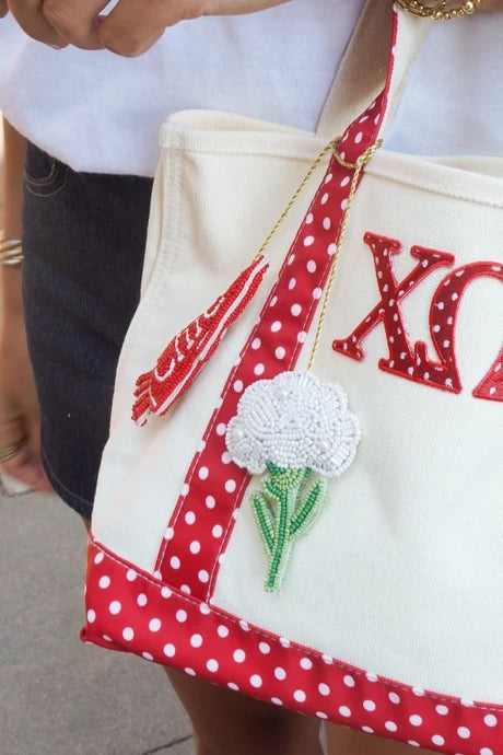 A cream canvas tote bag with red and white polka dot straps and trim, featuring a red sequined Greek letter 'XO' and a white flower applique. The bag is being held by a person wearing gold bracelets and a white shirt.