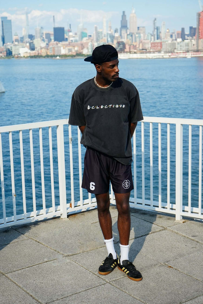 Young man wearing a black oversized classic friends logo t-shirt and shorts, standing by a waterfront with city skyline in the background
