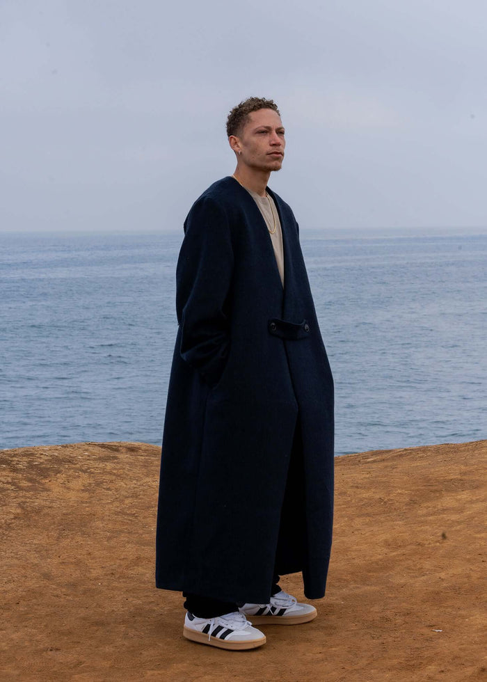 Man wearing a navy Collarless Long Coat by Éclectique Kanso, standing by the ocean.