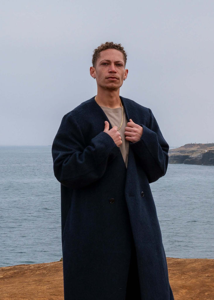 Model wearing a Collarless Long Coat Navy by Éclectique Kanso by the seaside with a calm ocean backdrop.