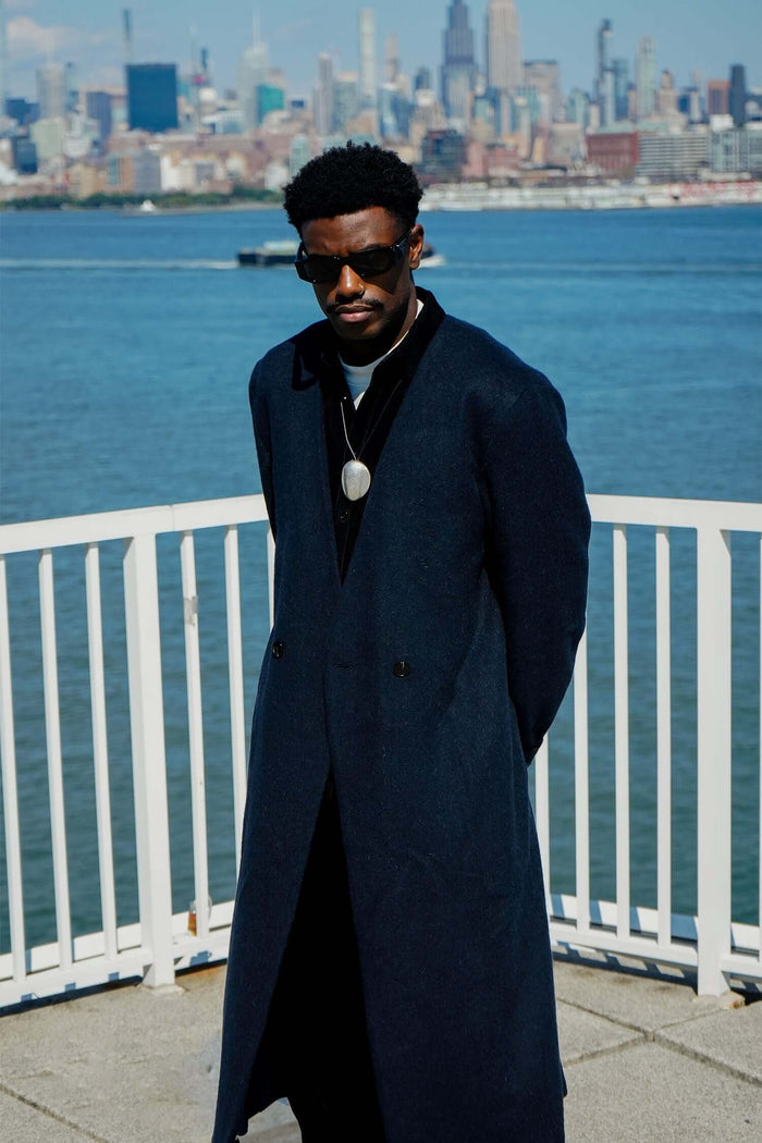 Man wearing a stylish Kanso Collarless Long Coat by Éclectique, standing by the waterfront with a city skyline in the background.