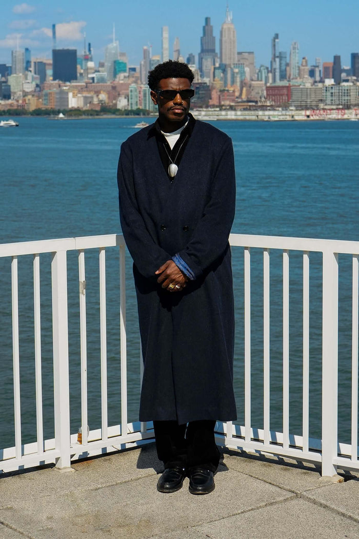 Model wearing the Kanso Collarless Long Coat with city skyline and waterfront in the background.