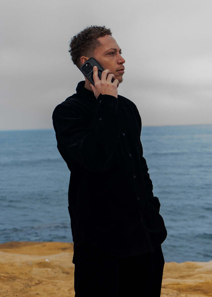 A man wearing a black corduroy button-up shirt, talking on a phone by the ocean.