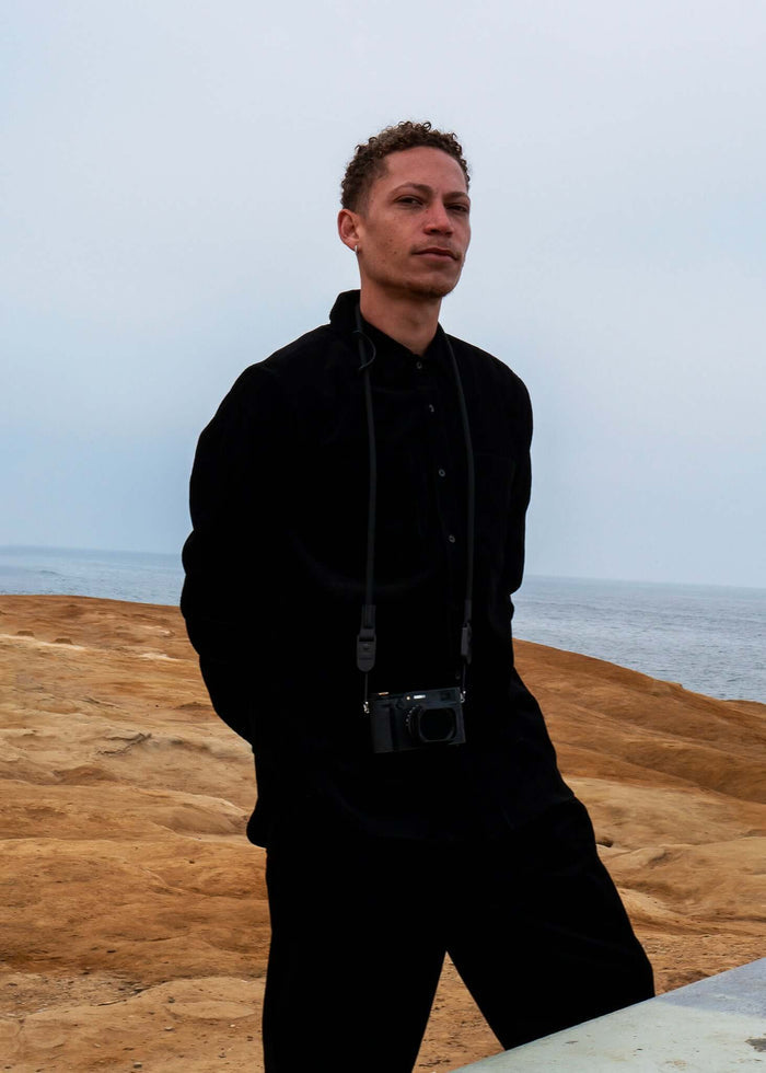 Man in black corduroy button-up shirt standing by water with a camera around his neck.
