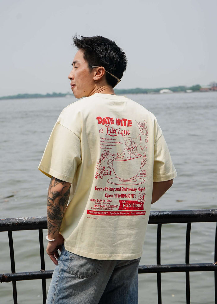 Model wearing a cream graphic T-shirt featuring Date Night artwork on the back, styled by Éclectique, near a waterfront.