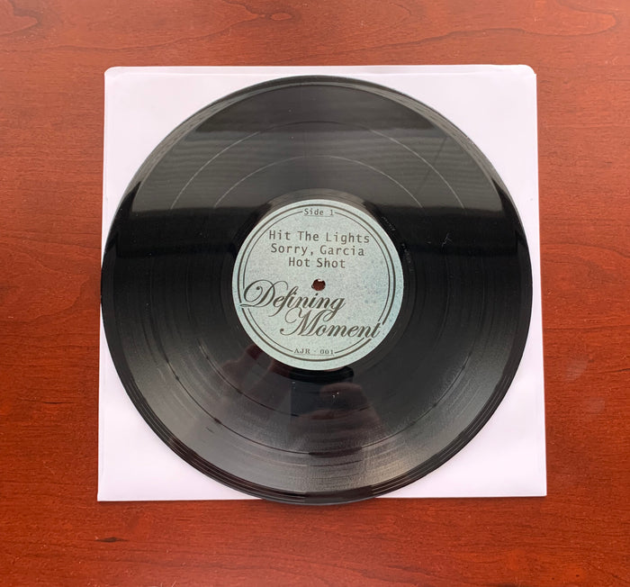 A black limited edition DZRP vinyl in a white paper sleeve rests on a reddish-brown wooden surface. The label lists Side 1: Hit The Lights, Sorry Garcia, Hot Shot, Defining Moment, with catalog number AJR-001.