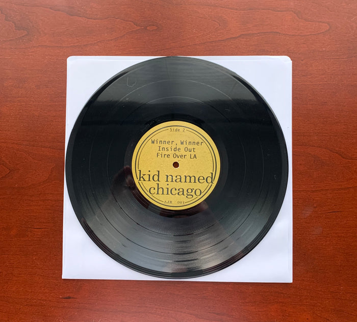 A limited edition DZRP vinyl, “kid named chicago,” sits on a white sleeve atop a reddish-brown wooden surface. Its label features songs such as “Winner, Winner,” inspired by the Tallahassee music scene and bands like Mayday Parade.