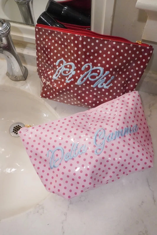Two polka dot cosmetic pouches on a bathroom sink countertop. The back pouch is dark red with white polka dots and silver script reading 'Pi Phi'.