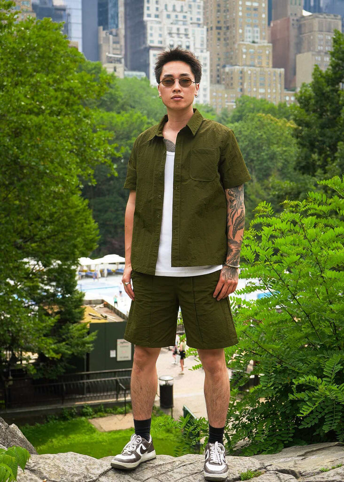 Model wearing olive green men's casual shorts and matching shirt in a park setting.