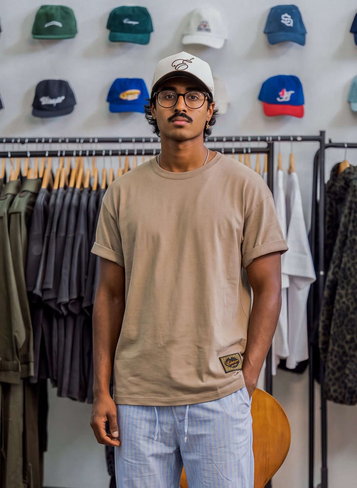 Model wearing Essential Cotton T-shirt in sand color, paired with a cap and light striped pants, in a trendy clothing store.