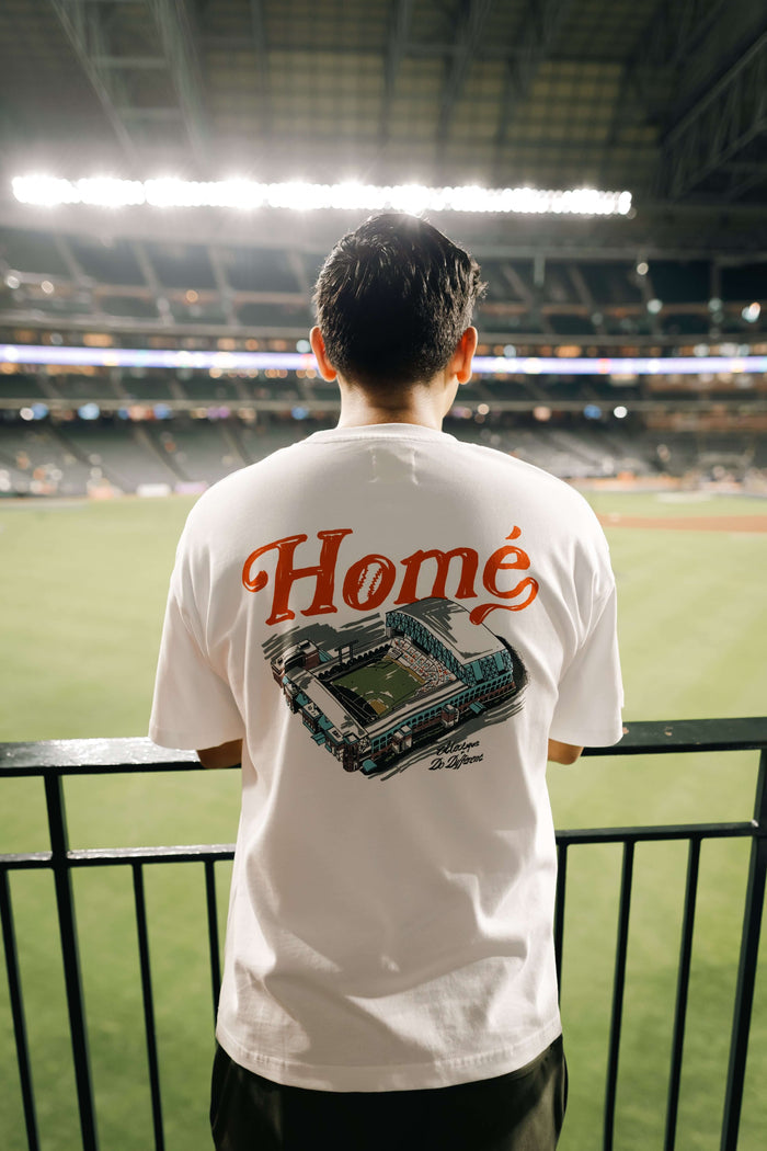 Home collaboration t-shirt worn by a person at a baseball game, featuring a graphic design on the back that says "Homé"