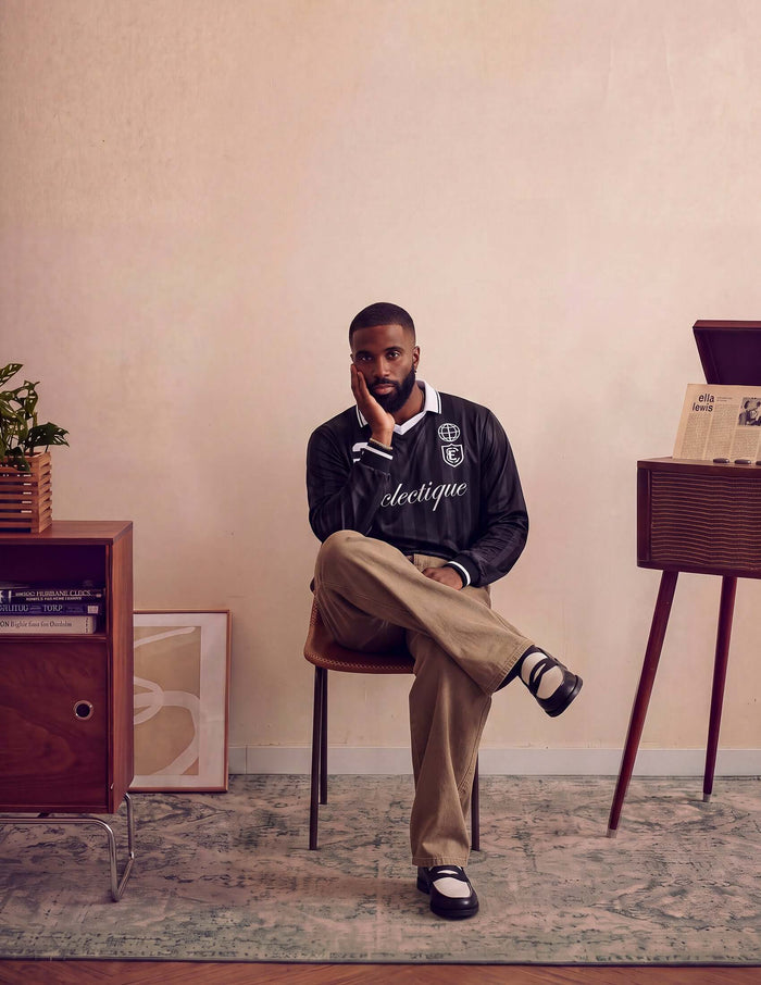 A man sitting on a chair wearing Kura Field Pleated Trousers and a stylish black jacket, in a modern interior setting.