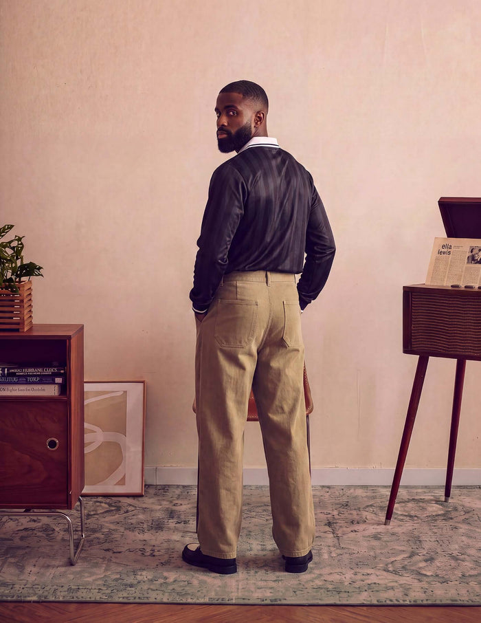 Model wearing Kura Field Pleated Trousers with a black shirt, facing back in a stylish interior setting.