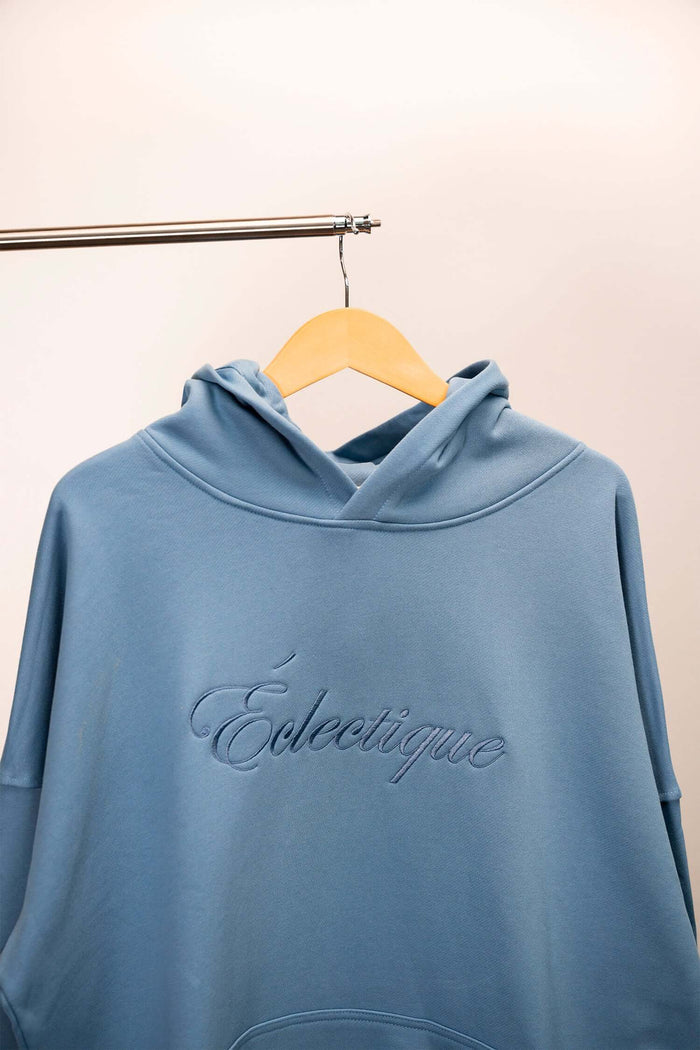 Lightweight terry hoodie with embroidered Éclectique script logo in light blue hanging on a wooden hanger.