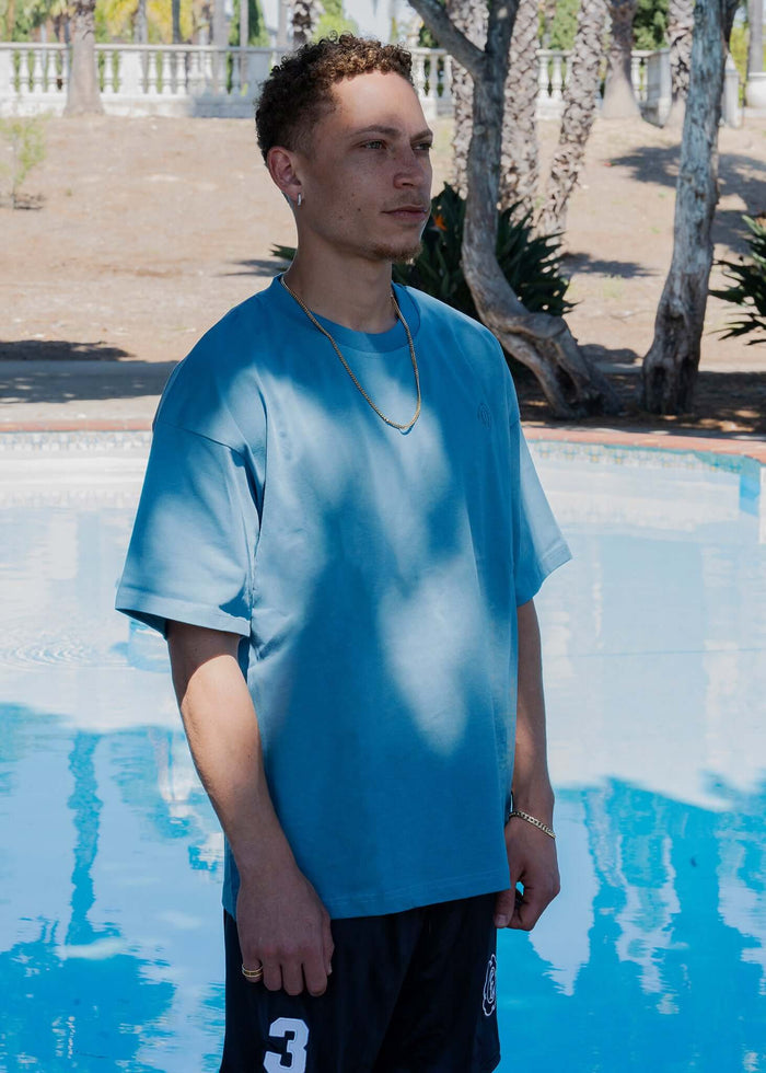 Person modeling a monogram embroidered cotton t-shirt in blue near a swimming pool.