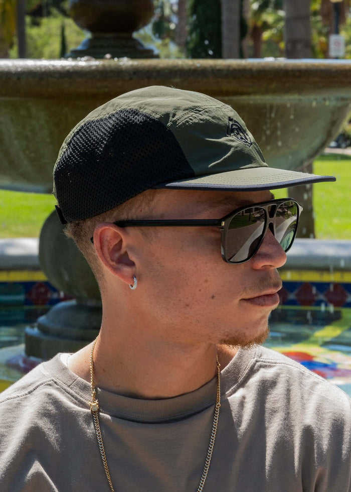 Monogram Performance Mesh Cap Olive worn by a man in sunglasses near a fountain in a park.