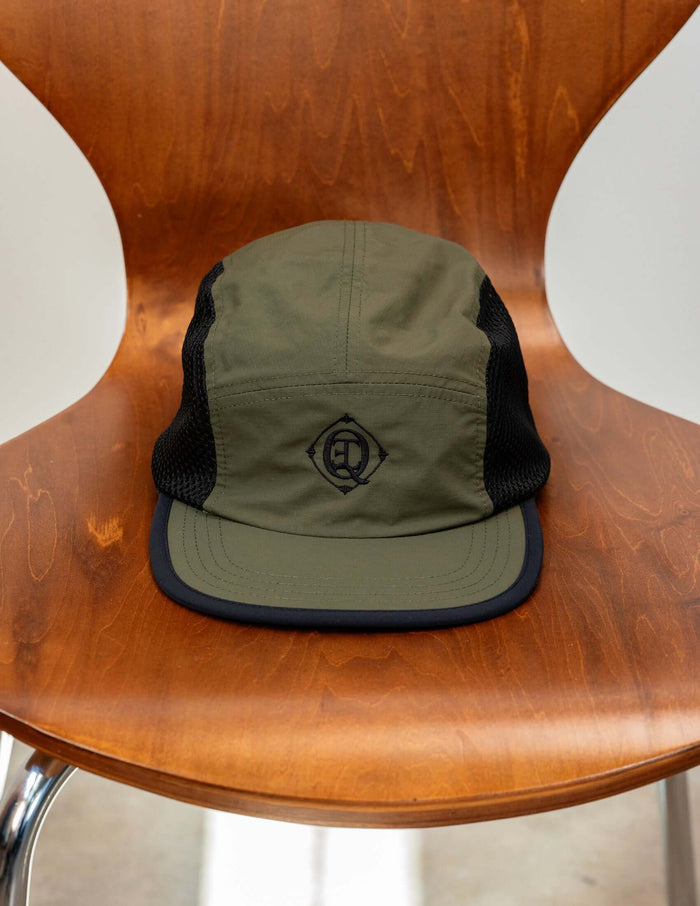 Monogram Performance Mesh Cap Olive displayed on a wooden chair, featuring a black logo and breathable mesh side panels.