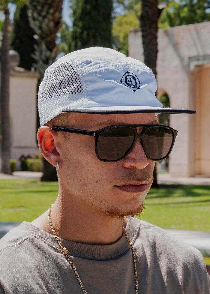 Man wearing a Monogram Performance Mesh Cap in white with sunglasses, outdoors surrounded by greenery.