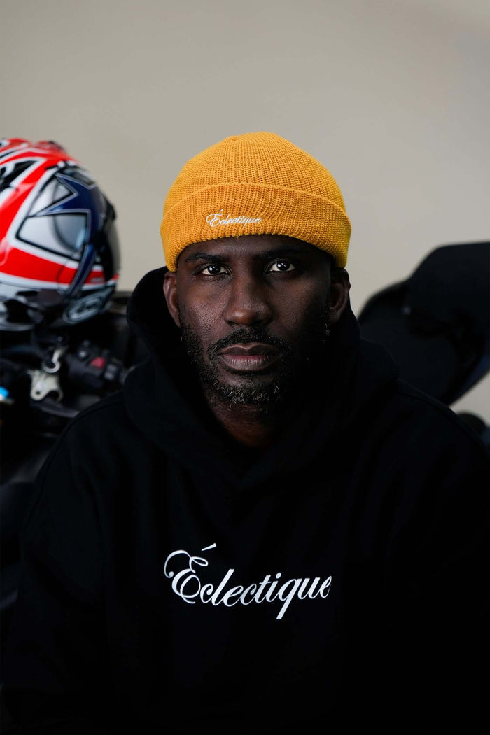 Mustard fisherman beanie worn by a model in a black hoodie with Éclectique branding, featuring a motorcycle helmet in the background.