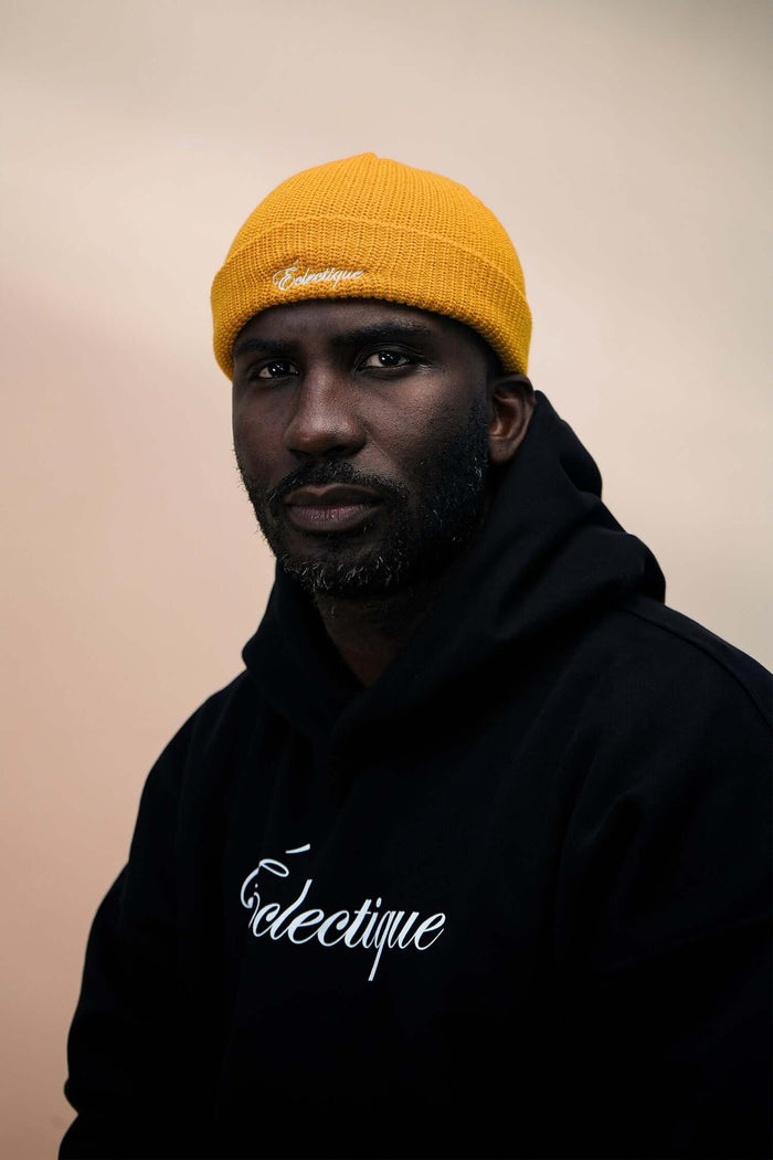 Portrait of a man wearing a mustard fisherman beanie and black hoodie with Éclectique logo.
