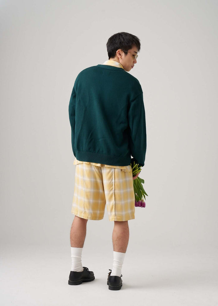 A man wearing an oversized green sweater and yellow ombre plaid shorts, holding tulips, seen from the back.