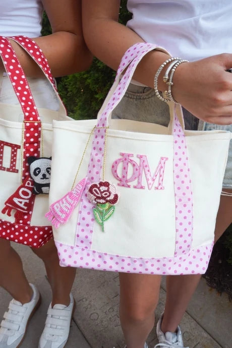 A cream canvas tote bag with pink polka-dot handles and trim, featuring pink monogrammed letters and a red rose patch, held by a person outdoors.