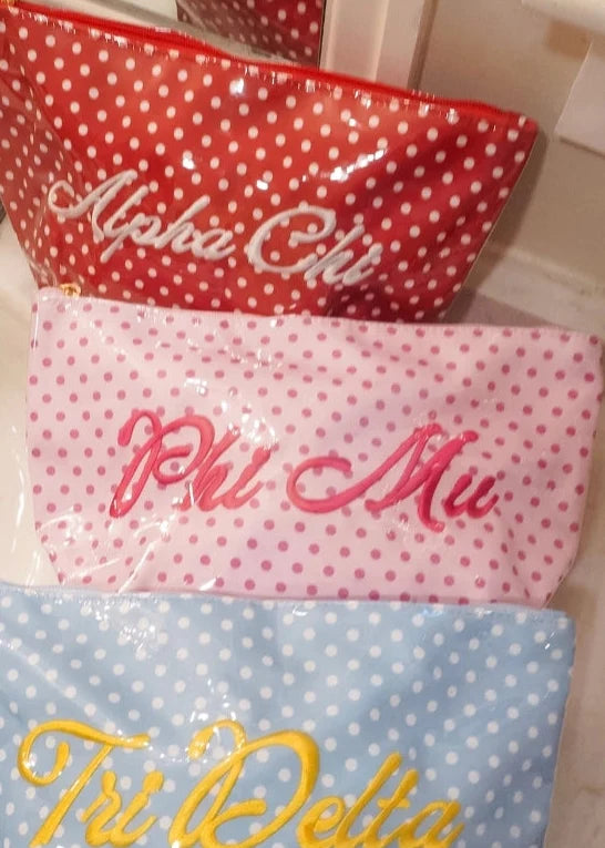 Three personalized zippered pouches with white polka dots are stacked on a bathroom sink. The top red pouch is labeled 'Alpha Chi' in white script, the middle pink pouch is labeled 'Phi Mu' in pink script, and the bottom blue pouch is labeled 'Tri Delta' in yellow script.