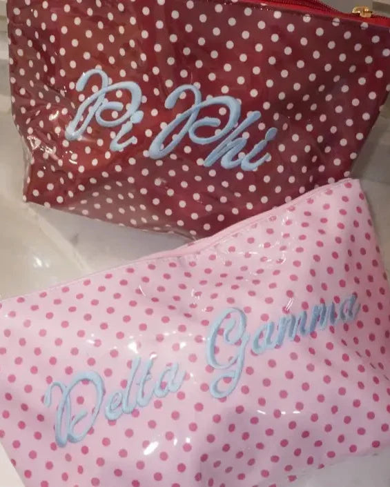 Two glossy vinyl pouches with white polka dots, one red and one pink, featuring blue cursive text reading 'Pi Phi' and 'Delta Gamma' respectively, resting on a bathroom sink.