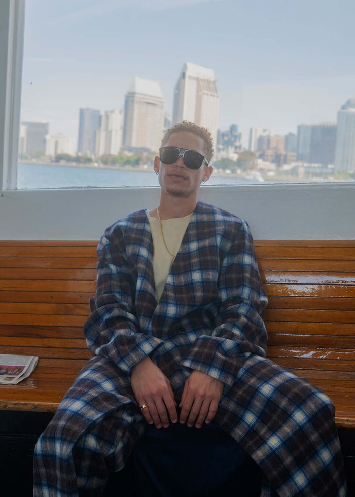 Model wearing a plaid collarless long coat sitting on a bench with city skyline in the background.