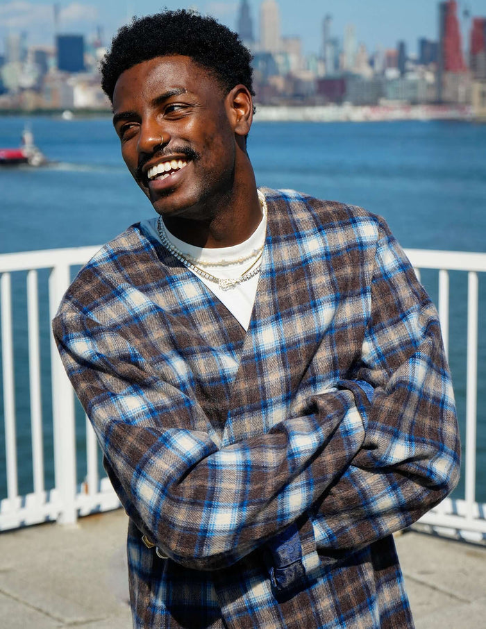 Model wearing a stylish plaid collarless long coat by Éclectique against a waterfront city backdrop.