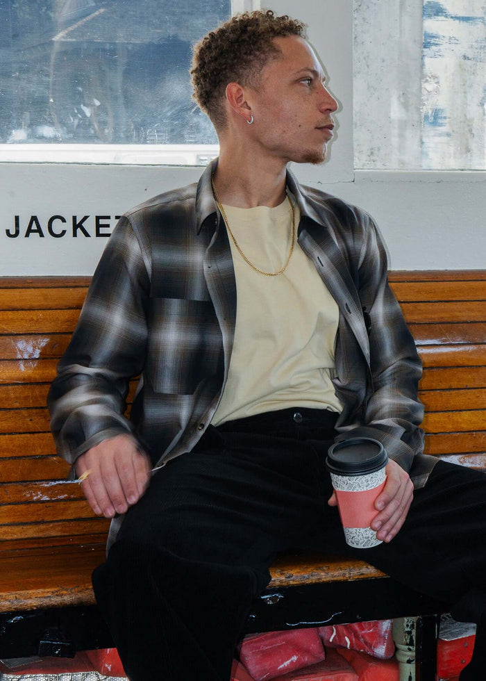 Man wearing a plaid cotton button-up shirt, sitting on a bench holding a coffee cup.