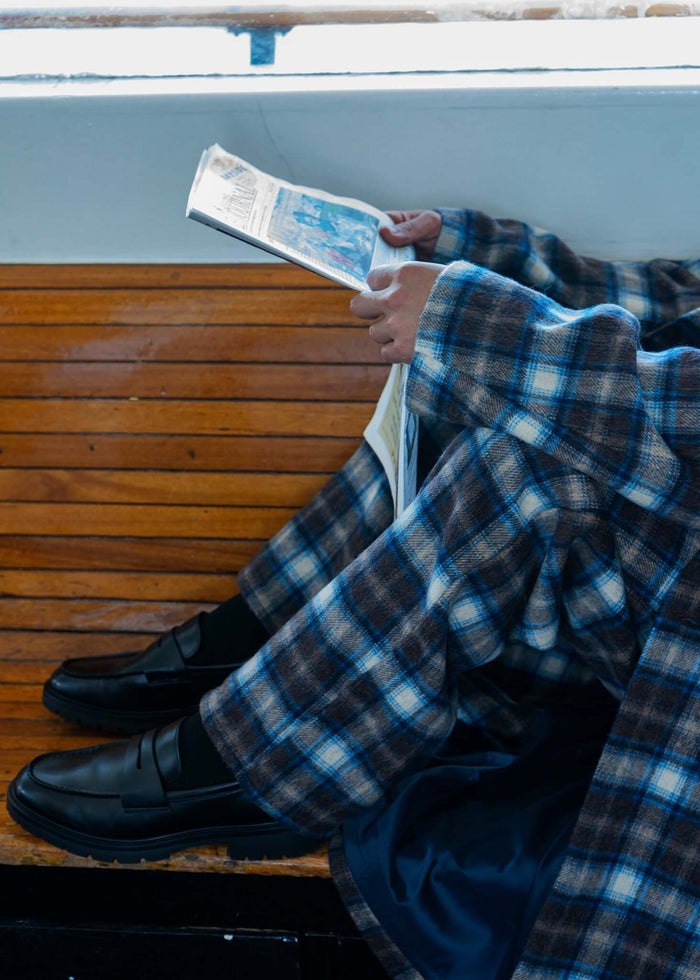 Model wearing plaid pleated trousers, sitting on a bench and reading a newspaper.