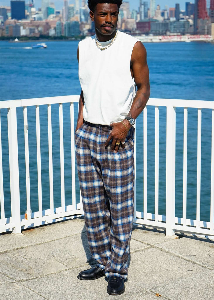 Man wearing plaid pleated trousers by the waterfront, showcasing a casual style against a city skyline.