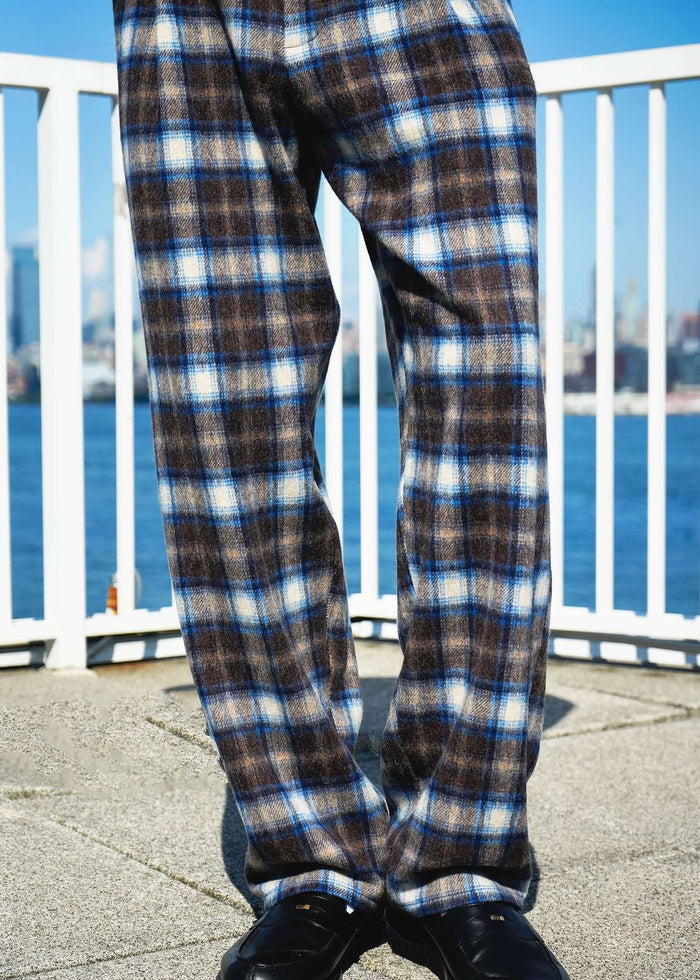 Plaid Pleated Trousers in brown and blue, displayed against a waterfront background.