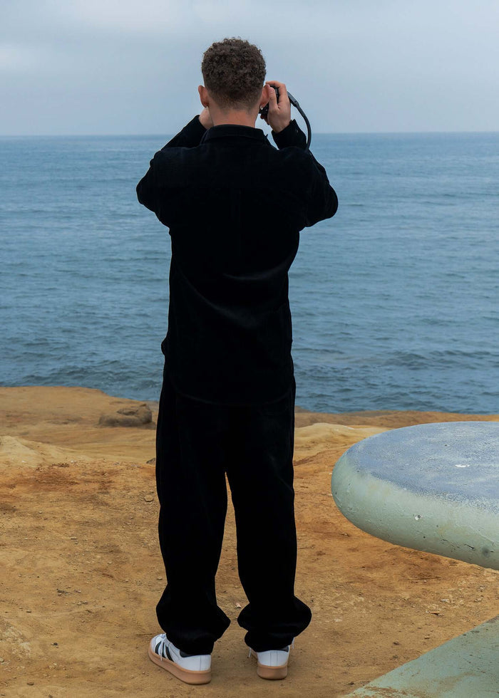 Person wearing Pleated Corduroy Trousers stands by the ocean, capturing a photo on a cloudy day.