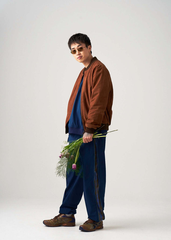 Model wearing brown bomber jacket, Reversible Windbreaker Pants, holding flowers in a studio setting.