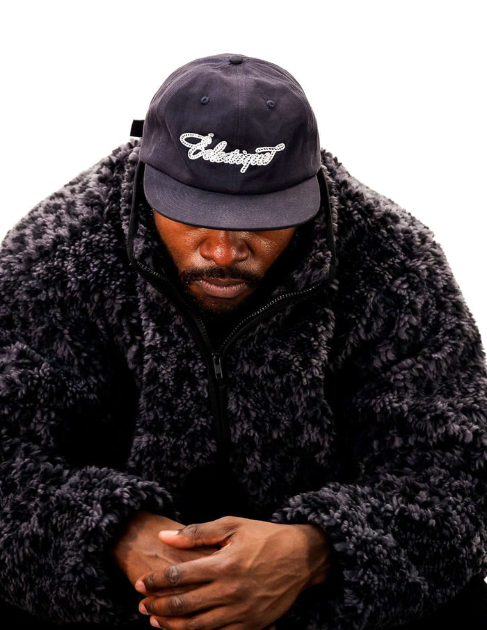 Rope script dad hat worn by a person in a black fur jacket, close-up view showcasing the embroidered design.
