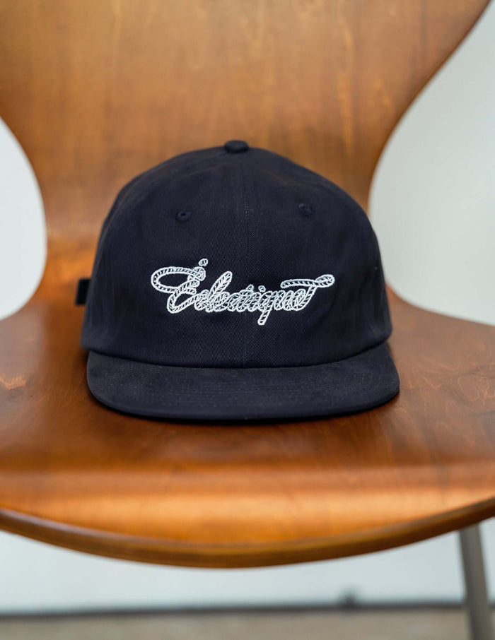 Rope script dad hat displayed on a wooden chair, featuring black cotton material and embroidered Éclectique script.