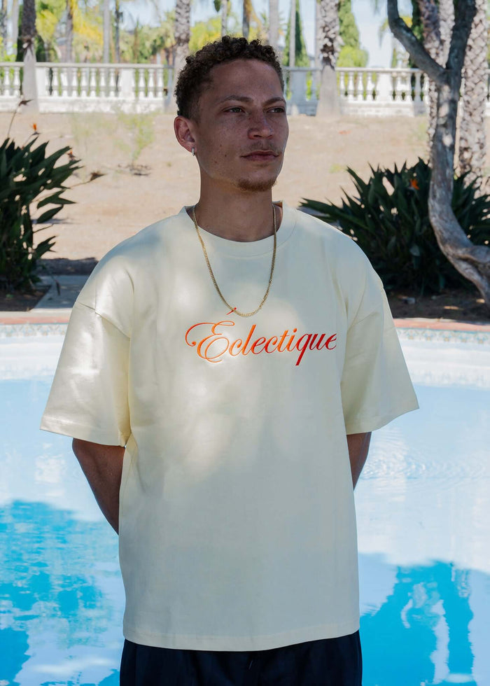 A model wearing a Script Logo Embroidered Cotton T-Shirt in French Vanilla by Éclectique, posed by a pool.