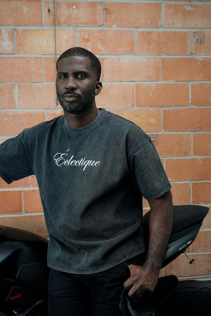 Man wearing a script logo t-shirt in acid-wash black, posing by a motorcycle against a brick wall.